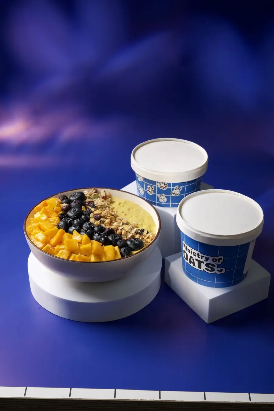 A mango and blueberry bowl beside two Ministry of Oats tubs.